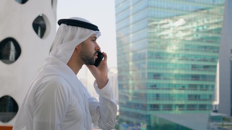 جهاز تقني حديث ومتطور، التحدث والتواصل مع الاهل والاصدقاء عبر الجوال، تصوير مقرب من الجانب لرجل عربي خليجي اماراتي يرتدي الكندورة والغترة يقوم بإجراء مكالمة هاتفية باستخدام الهاتف المحمول، الأبراج وناطحات السحاب في الامارات