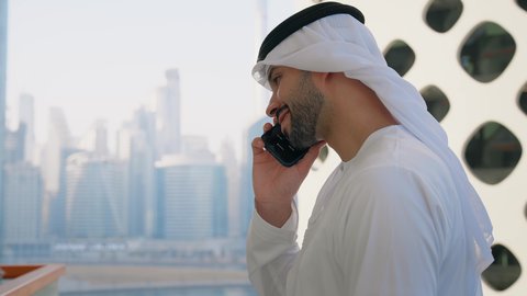 ايماءات السعادة والسرور، جهاز تقني حديث ومتطور، التحدث والتواصل مع الاهل والاصدقاء عبر الجوال، تصوير مقرب من الجانب لرجل عربي خليجي اماراتي يرتدي الكندورة والغترة يقوم بإجراء مكالمة هاتفية باستخدام الهاتف المحمول، الأبراج وناطحات السحاب في الامارات