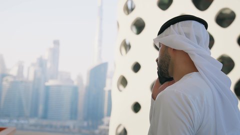 Expressions of happiness and pleasure, a modern and advanced technical device, talking and communicating with family and friends via mobile phone, a close-up shot from the side of an Arab Gulf Emirati man wearing a kandura and ghutra making a phone call using a mobile phone, towers and skyscrapers in the Emirates