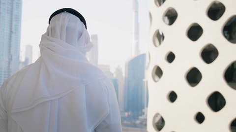 التواصل مع الاهل والاصدقاء عبر الجوال، استخدام جهاز تقني حديث ومتطور، تصوير مقرب من الخلف لرجل عربي خليجي اماراتي يرتدي الكندورة والغترة يقوم بإجراء مكالمة هاتفية باستخدام الهاتف المحمول، الأبراج وناطحات السحاب في الامارات