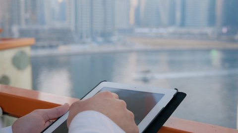 إنجاز الأعمال عن بعد عبر الانترنت، جهاز تقني حديث ومتطور، التواصل مع الاهل والاصدقاء عبر الجهاز اللوحي، تصوير مقرب لأيدي رجل عربي خليجي اماراتي يقوم باستخدام التابليت المحمول، خلفية الأبراج وناطحات السحاب في الامارات