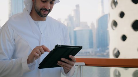 تنظيم العمل عن بعد عبر الانترنت، جهاز تقني حديث ومتطور، التواصل مع الاهل والاصدقاء عبر الجهاز اللوحي، تصوير مقرب لرجل عربي خليجي اماراتي مبتسم يقوم باستخدام التابليت المحمول، خلفية الأبراج وناطحات السحاب في الامارات نهارا