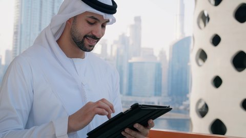 تصفح مواقع التواصل الاجتماعي، العمل عن بعد عبر الانترنت، جهاز تقني حديث ومتطور، التواصل مع الاهل والاصدقاء عبر الجهاز اللوحي، تصوير مقرب لرجل عربي خليجي اماراتي مبتسم يقوم باستخدام التابليت المحمول، خلفية الأبراج وناطحات السحاب في الامارات نهارا