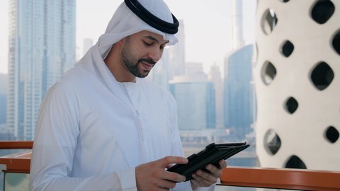 رفع قبضة اليد بتعابير الفوز والسرور، تنظيم العمل عن بعد عبر الانترنت، جهاز تقني حديث ومتطور، التواصل مع الاهل والاصدقاء عبر الجهاز اللوحي، تصوير مقرب لرجل عربي خليجي اماراتي مبتسم يرتدي الكندورة والغترة يقوم باستخدام التابليت المحمول، خلفية الأبراج في الامارات