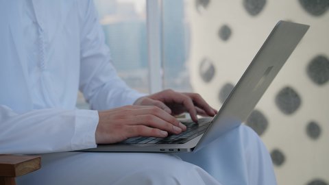 متابعة العمل عن بعد عبر الانترنت، جهاز تقني حديث ومتطور، تصوير مقرب لرجل عربي خليجي اماراتي يرتدي الكندورة والغترة يقوم باستخدام الحاسوب المحمول بتعابير السرور، خلفية الأبراج وناطحات السحاب في الامارات