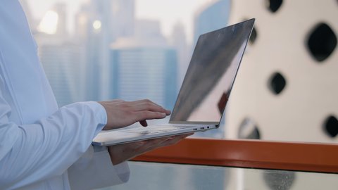 Working remotely online, a modern and advanced technical device, a close-up of an Emirati Gulf Arab man wearing a kandura and ghutra using a laptop with an expression of pleasure, a background of towers and skyscrapers in the Emirates