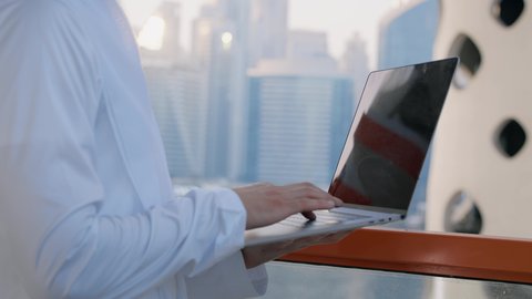استخدام التكنولوجيا والتقنيات الحديثة، مفهوم إنجاز العمل عن بعد عبر جهاز اللابتوب، تصوير مقرب لرجل عربي خليجي اماراتي يرتدي الكندورة يقوم باستخدام الحاسوب المحمول، خلفية الأبراج وناطحات السحاب في الامارات