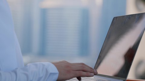 Using modern technology and techniques, the concept of completing work remotely via laptop, a close-up of an Emirati Gulf Arab man wearing a kandura using a laptop, a background of towers and skyscrapers in the Emirates