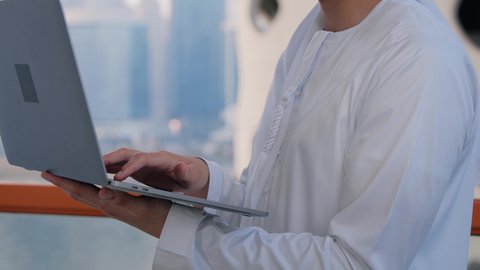 إنجاز العمل عن بعد عبر جهاز اللابتوب، استخدام التكنولوجيا والتقنيات الحديثة، تصوير مقرب لرجل عربي خليجي اماراتي يرتدي الكندورة يقوم باستخدام الحاسوب المحمول، خلفية الأبراج وناطحات السحاب في الامارات