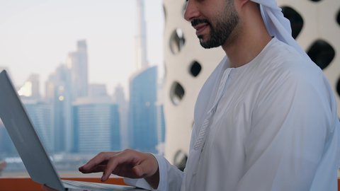 جلسة هادئة في الخارج، تنظيم العمل عن بعد عبر الانترنت، جهاز تقني حديث ومتطور، تصوير مقرب لرجل عربي خليجي اماراتي يرتدي الكندورة والغترة يقوم باستخدام الحاسوب المحمول بتعابير السرور، خلفية الأبراج وناطحات السحاب في الامارات