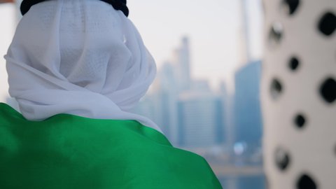 حب الوطن والاعتزاز به، الاحتفال بذكرى تأسيس الاتحاد 2 ديسمبر في الطريق، يوم العلم 3 نوفمبر، اليوم الوطني الاماراتي 1971، تصوير من الخلف لرجل عربي خليجي اماراتي يرتدي الكندورة والغترة يضع علم الامارات العربية المتحدة على كتفيه في النهار