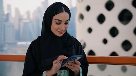 Browsing social media applications and programs, chatting and exchanging conversations and news with family and friends, a close-up of an Emirati Gulf Arab woman wearing an abaya and hijab holding a mobile phone, using a modern and advanced technical device, buildings and facilities overlooking the sea in the Emirates