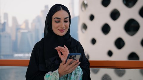 الأبراج والمنشآت المطلة على البحر في الامارات، تصفح التطبيقات وبرامج التواصل الاجتماعي، الدردشة و تبادل الأحاديث والأخبار مع الأهل والأصدقاء، تصوير مقرب لامراة عربية خليجية اماراتية ترتدي العباءة والحجاب تمسك بيدها الهاتف المحمول، استخدام جهاز تقني حديث ومتطور