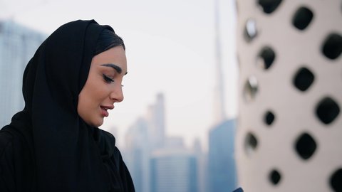 ايماءات السعادة والسرور، اجراء مكالمة فيديو عبر جهاز الجوال، التواصل وتبادل الأحاديث والأخبار مع الأهل والأصدقاء، تصوير مقرب لامراة عربية خليجية اماراتية ترتدي العباءة والحجاب تمسك بيدها الهاتف المحمول، استخدام جهاز تقني حديث ومتطور، قضاء وقت ممتع في الخارج