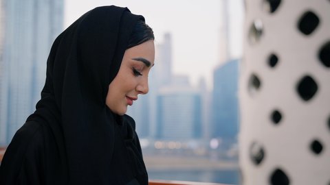 قضاء وقت ممتع في الخارج، ايماءات السعادة والسرور، اجراء مكالمة فيديو عبر جهاز الجوال، التواصل وتبادل الأحاديث والأخبار مع الأهل والأصدقاء، تصوير مقرب لامراة عربية خليجية اماراتية ترتدي العباءة والحجاب تمسك بيدها الهاتف المحمول، استخدام جهاز تقني حديث ومتطور