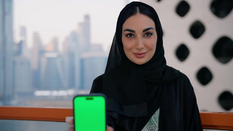 النظر إلى الكاميرا بايماءات السعادة والسرور، عرض شاشة خضراء فارغة عبر الجوال، كروما، قضاء وقت ممتع في الخارج، تصوير مقرب لامراة عربية خليجية اماراتية ترتدي العباءة والحجاب تمسك بيدها الهاتف المحمول، استخدام جهاز تقني حديث ومتطور