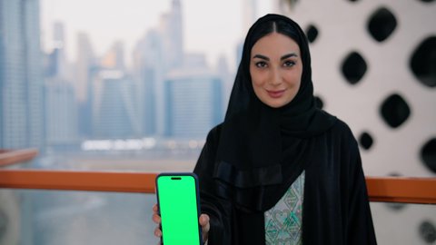 عرض شاشة خضراء فارغة عبر الجوال، كروما، قضاء وقت ممتع في الخارج، النظر إلى الكاميرا بايماءات السعادة والسرور، تصوير مقرب لامراة عربية خليجية اماراتية ترتدي العباءة والحجاب تمسك بيدها الهاتف المحمول، استخدام جهاز تقني حديث ومتطور