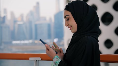 الأبراج والمنشآت المطلة على البحر في الامارات، تصفح التطبيقات وبرامج التواصل الاجتماعي، الدردشة و تبادل الأحاديث والأخبار مع الأهل والأصدقاء، تصوير مقرب لامراة عربية خليجية اماراتية ترتدي العباءة والحجاب تمسك بيدها الهاتف المحمول، استخدام جهاز تقني حديث ومتطور