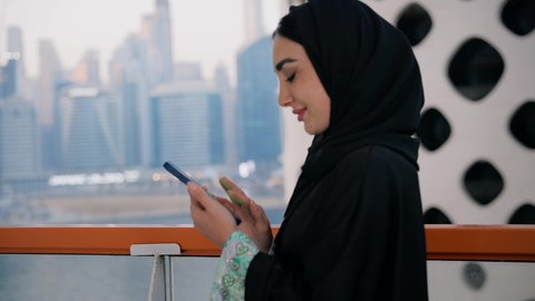 تصفح التطبيقات وبرامج التواصل الاجتماعي، الدردشة و تبادل الأحاديث والأخبار مع الأهل والأصدقاء، تصوير مقرب لامراة عربية خليجية اماراتية ترتدي العباءة والحجاب تمسك بيدها الهاتف المحمول، استخدام جهاز تقني حديث ومتطور، المباني والمنشآت المطلة على البحر في الامارات