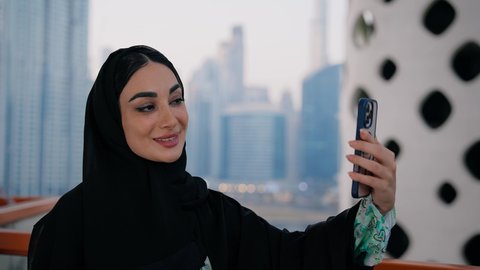 توثيق اللحظات التذكارية السعيدة، استخدام جهاز تقني حديث ومتطور لتخليد الذكريات ، التقاط الصور الشخصية باستخدام الهاتف المحمول، تصوير مقرب لامراة عربية خليجية اماراتية ترتدي العباءة والحجاب تأخذ صورة سيلفي بإيماءات السعادة والسرور