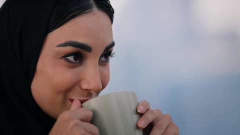 Having a good time outside, enjoying hot drinks, gestures of pleasure and delight in sipping a favorite drink, eating warm winter herbs, close-up of an Emirati woman wearing an abaya and hijab drinking coffee