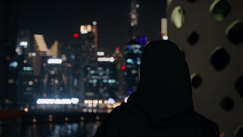 قضاء وقت ممتع في الخارج, تصوير مقرب لامراة اماراتية ترتدي العباءة السوداء والحجاب تقوم بتأمل الأبراج وناطحات السحاب المضاءة في وقت الليل في الامارات، الأناقة والاهتمام بالمظهر الخارجي