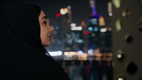 تأمل الأبراج وناطحات السحاب المضاءة في وقت الليل في الامارات، تناول الأعشاب الشتوية الدافئة، قضاء وقت ممتع في الخارج، الاستمتاع بالمشروبات الساخنة، ايماءات المتعة والتلذذ باحتساء المشروب المفضل, تصوير مقرب لامراة اماراتية ترتدي العباءة والحجاب تقوم بشرب القهوة