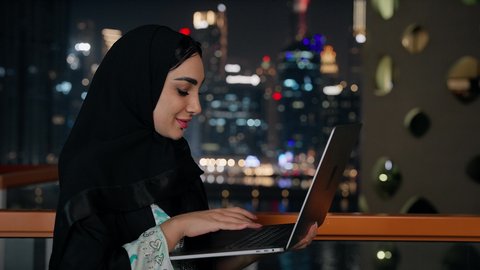 ادارة الأعمال وانجاز المهام المطلوبة، العمل عن بعد، مهنة ووظيفة نسائية، تصوير مقرب لإمرأة عربية خليجية اماراتية محجبة ترتدي عباءة ملونة تستخدم جهاز اللابتوب، استخدام جهاز تقني حديث ومتطور، الأبراج والمنشآت المضاءة في وقت الليل في الامارات