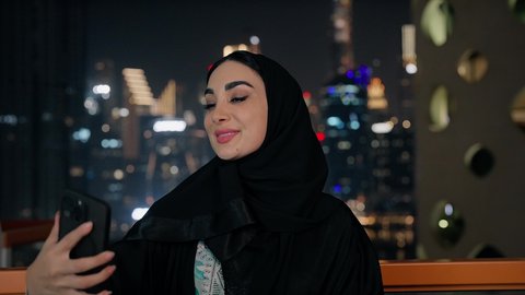 Documenting happy memorable moments, using a modern and advanced technical device to immortalize memories, taking personal photos using a mobile phone, a close-up of an Emirati Gulf Arab woman wearing an abaya and hijab taking a selfie with gestures of happiness and pleasure