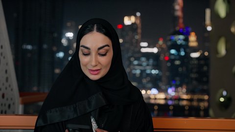 التواصل وتبادل الأحاديث والأخبار مع الأهل والأصدقاء، قضاء وقت ممتع في الخارج، ايماءات السعادة والسرور، اجراء مكالمة هاتفية عبر جهاز الجوال، تصوير مقرب لامراة عربية خليجية اماراتية ترتدي العباءة والحجاب تمسك بيدها الهاتف المحمول، استخدام جهاز تقني حديث ومتطور
