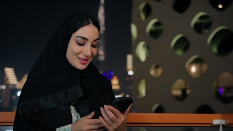 ايماءات السعادة والسرور، اجراء مكالمة فيديو عبر جهاز الجوال، التواصل وتبادل الأحاديث والأخبار مع الأهل والأصدقاء، تصوير مقرب لامراة عربية خليجية اماراتية ترتدي العباءة والحجاب تمسك بيدها الهاتف المحمول، استخدام جهاز تقني حديث ومتطور، قضاء وقت ممتع في الخارج
