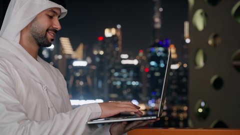 جلسة هادئة في الخارج، تنظيم العمل عن بعد عبر الانترنت، جهاز تقني حديث ومتطور، تصوير مقرب لرجل عربي خليجي اماراتي يرتدي الكندورة والغترة يقوم باستخدام الحاسوب المحمول بتعابير السرور، خلفية الأبراج وناطحات السحاب في الامارات