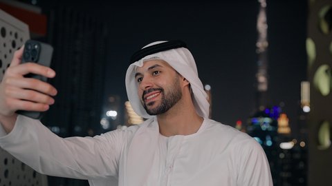 استخدام جهاز تقني حديث ومتطور في تخليد الذكريات، التقاط الصور الشخصية باستخدام الجوال، توثيق اللحظات السعيدة، تصوير مقرب لرجل عربي خليجي اماراتي مبتسم يرتدي الكندورة والغترة البيضاء يمسك الهاتف المحمول ويأخذ صورة سيلفي تذكارية بإيماءات السرور، خلفية الأبراج ليلا