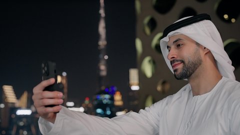 Documenting happy moments, taking selfies with a mobile phone, using a modern and advanced technical device to immortalize memories, a close-up of a smiling Emirati Gulf Arab man wearing a kandura and a white ghutra holding a mobile phone and taking a commemorative selfie with gestures of pleasure