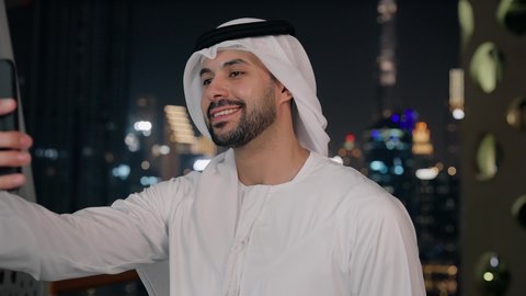 استخدام جهاز تقني حديث ومتطور، التواصل مع الاهل والاصدقاء عبر الجوال، تصوير مقرب لرجل عربي خليجي اماراتي مبتسم يرتدي الكندورة والغترة يقوم بإجراء مكالمة فيديو باستخدام الهاتف المحمول بإيماءات السرور والسعادة، خلفية الأبراج المضاءة ليلا