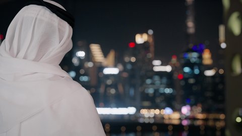 التواصل مع الاهل والاصدقاء عبر الجوال، استخدام جهاز تقني حديث ومتطور، تصوير مقرب من الخلف لرجل عربي خليجي اماراتي يرتدي الكندورة والغترة يقوم بإجراء مكالمة هاتفية باستخدام الهاتف المحمول، الأبراج وناطحات السحاب في الامارات