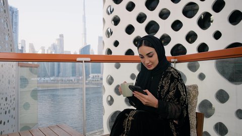 تصفح التطبيقات وبرامج التواصل الاجتماعي، تبادل الأحاديث والأخبار مع الأهل والأصدقاء،  تصوير مقرب لامراة اماراتية ترتدي العباءة والحجاب تمسك بيدها الهاتف المحمول وتشرب القهوة، استخدام جهاز تقني حديث ومتطور، احتساء المشروب المفضل في المقهى