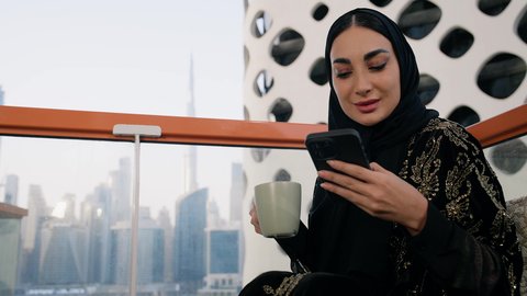 احتساء المشروب المفضل في المقهى، تصفح التطبيقات وبرامج التواصل الاجتماعي، تبادل الأحاديث والأخبار مع الأهل والأصدقاء،  تصوير مقرب لامراة اماراتية ترتدي العباءة والحجاب تمسك بيدها الهاتف المحمول وتشرب القهوة، استخدام جهاز تقني حديث ومتطور