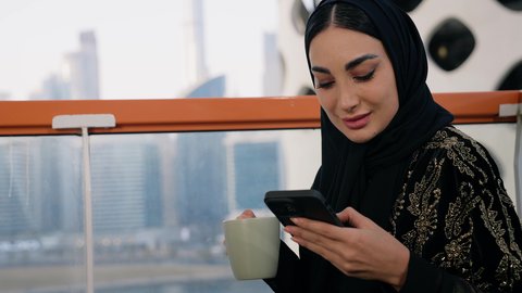 احتساء المشروب المفضل في المقهى، تصفح التطبيقات وبرامج التواصل الاجتماعي، تبادل الأحاديث والأخبار مع الأهل والأصدقاء،  تصوير مقرب لامراة اماراتية ترتدي العباءة والحجاب تمسك بيدها الهاتف المحمول وتشرب القهوة، استخدام جهاز تقني حديث ومتطور