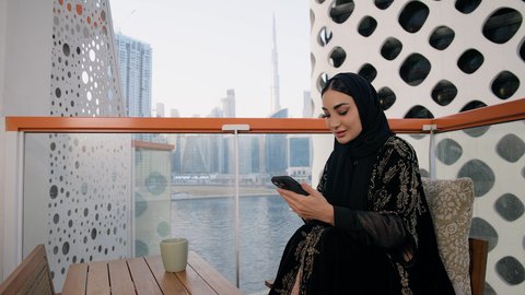 احتساء المشروب المفضل في المقهى، تصفح التطبيقات وبرامج التواصل الاجتماعي، تبادل الأحاديث والأخبار مع الأهل والأصدقاء،  تصوير مقرب لامراة اماراتية ترتدي العباءة والحجاب تمسك بيدها الهاتف المحمول وتشرب القهوة، استخدام جهاز تقني حديث ومتطور