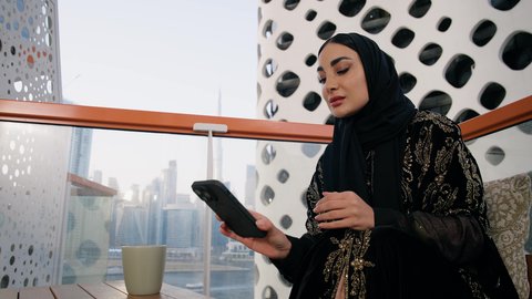 تصفح التطبيقات وبرامج التواصل الاجتماعي، تبادل الأحاديث والأخبار مع الأهل والأصدقاء،  تصوير مقرب لامراة اماراتية ترتدي العباءة والحجاب تمسك بيدها الهاتف المحمول وتشرب القهوة، استخدام جهاز تقني حديث ومتطور، احتساء المشروب المفضل في المقهى