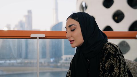 التواصل وتبادل الأحاديث والأخبار مع الأهل والأصدقاء،  تصوير مقرب لامراة اماراتية ترتدي العباءة والحجاب تمسك بيدها الهاتف المحمول وتشرب القهوة، استخدام جهاز تقني حديث ومتطور، احتساء المشروب المفضل في المقهى، اجراء مكالمة هاتفية عبر الجوال