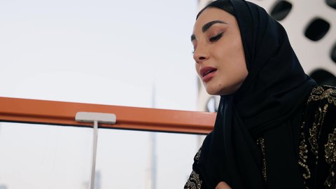 اجراء مكالمة هاتفية عبر جهاز الجوال، التواصل وتبادل الأحاديث والأخبار مع الأهل والأصدقاء،  تصوير مقرب لامراة اماراتية ترتدي العباءة والحجاب تمسك بيدها الهاتف المحمول وتشرب القهوة، استخدام جهاز تقني حديث ومتطور، احتساء المشروب المفضل في المقهى