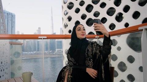 توثيق اللحظات التذكارية السعيدة، استخدام جهاز تقني حديث ومتطور لتخليد الذكريات ، التقاط الصور الشخصية باستخدام الهاتف المحمول، تصوير مقرب لامراة اماراتية ترتدي العباءة والحجاب تأخذ صورة سيلفي بإيماءات السعادة والسرور