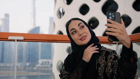 استخدام جهاز تقني حديث ومتطور لتخليد الذكريات ، التقاط الصور الشخصية باستخدام الهاتف المحمول، تصوير مقرب لامراة اماراتية ترتدي العباءة والحجاب تأخذ صورة سيلفي بإيماءات السعادة والسرور، توثيق اللحظات التذكارية السعيدة