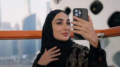 استخدام جهاز تقني حديث ومتطور لتخليد الذكريات ، التقاط الصور الشخصية باستخدام الهاتف المحمول، تصوير مقرب لامراة اماراتية ترتدي العباءة والحجاب تأخذ صورة سيلفي بإيماءات السعادة والسرور، توثيق اللحظات التذكارية السعيدة