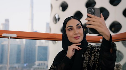 توثيق اللحظات التذكارية السعيدة، استخدام جهاز تقني حديث ومتطور لتخليد الذكريات ، التقاط الصور الشخصية باستخدام الهاتف المحمول، تصوير مقرب لامراة اماراتية ترتدي العباءة والحجاب تأخذ صورة سيلفي بإيماءات السعادة والسرور