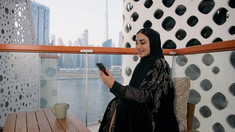 اجراء مكالمة هاتفية عبر جهاز الجوال، التواصل وتبادل الأحاديث والأخبار مع الأهل والأصدقاء،  تصوير مقرب لامراة اماراتية ترتدي العباءة والحجاب تمسك بيدها الهاتف المحمول وتشرب القهوة، استخدام جهاز تقني حديث ومتطور، احتساء المشروب المفضل في المقهى
