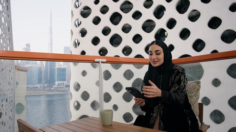اجراء مكالمة هاتفية عبر جهاز الجوال، التواصل وتبادل الأحاديث والأخبار مع الأهل والأصدقاء،  تصوير مقرب لامراة اماراتية ترتدي العباءة والحجاب تمسك بيدها الهاتف المحمول وتشرب القهوة، استخدام جهاز تقني حديث ومتطور، احتساء المشروب المفضل في المقهى