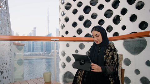 احتساء المشروب المفضل في الكافيه، ادارة الأعمال وانجاز المهام المطلوبة، العمل عن بعد، مهنة ووظيفة نسائية، تصوير مقرب لإمرأة عربية خليجية اماراتية محجبة ترتدي عباءة ملونة تستخدم الجهاز اللوحي وتشرب القهوة، استخدام جهاز تقني حديث ومتطور